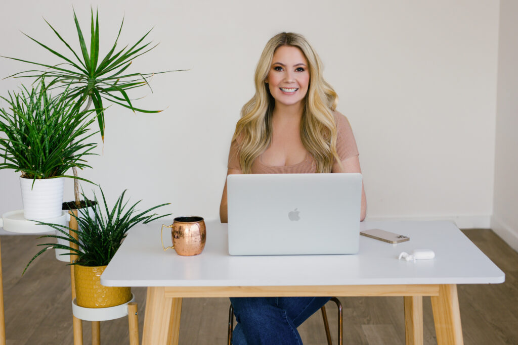 Returning client Elizabeth in a pinkish tan ribbed shirt with a square neck sitting at a white desk with an apple macbook pro open, iphone, airpods, and a copper coffee mug. 